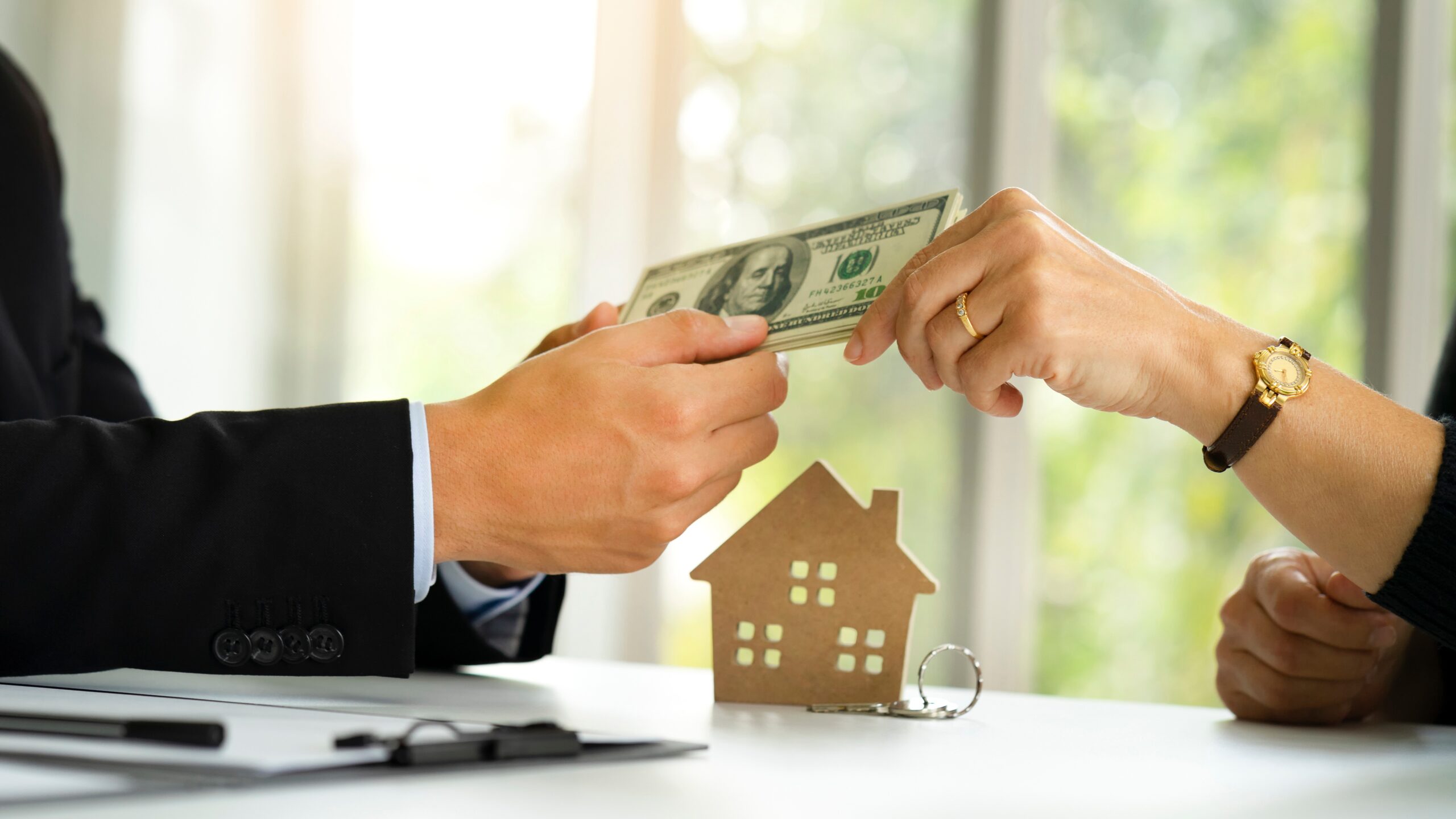 Image of a man in a suit handing money to a woman seated across the table from him with a miniature model of a house under their hands